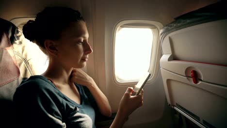 Tourist-woman-sitting-near-airplane-window-at-sunset-and-using-mobile-phone-during-flight