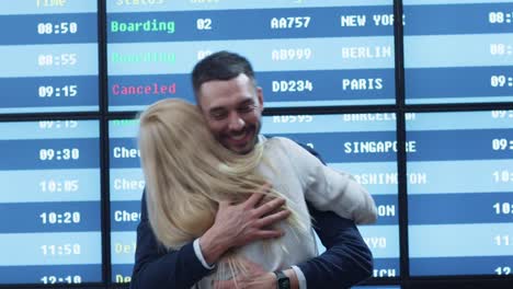 Man-and-Woman-meeting-in-the-Airport-Arrival-Hall-next-to-Information-Board.-People-are-Hugging-each-other-and-Smiling.