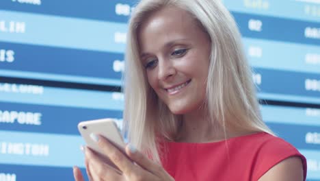 Atractivo-sonriente-rubia-mujer-usando-teléfono-móvil-junto-al-tablero-de-la-información-en-el-aeropuerto.