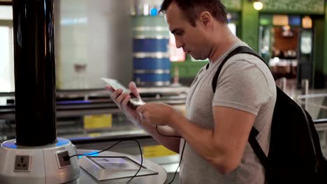 Hübscher-junger-Mann-im-Flughafen-terminal.-Stand-in-der-Nähe-laden-Stand-Pluging-in-smartphone