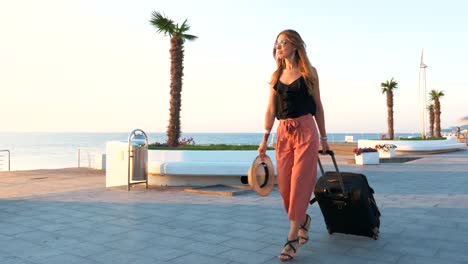 Young-business-lady-walking-with-suitcase-at-the-seafront-during-sunset