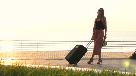 Young-business-lady-with-suitcase-at-the-seafront-during-sunset,-dolly