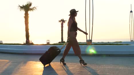 Young-business-lady-walking-with-suitcase-at-the-seafront-during-sunset