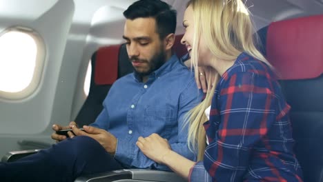 En-un-tablero-de-avión-comercial-hermosa-rubia-joven-con-macho-hispano-ver-Videos-en-el-teléfono-inteligente-y-sonrisa.-El-sol-brilla-a-través-de-la-ventana-del-avión.