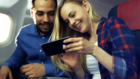 Bei-einem-Board-von-kommerziellen-Flugzeug-schöne-junge-Blondine-mit-schönen-hispanische-männlich-nutzen-Smartphone-und-lachen.-Durch-Flugzeug-Fenster-scheint-die-Sonne.