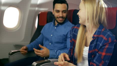 On-a-Commercial-Flight-Beautiful-Blonde-Female-Teaches-Handsome-Hispanic-Man-how-to-Play-Guitar.-Airplane-Is-New,-Sun-Shines-Through-Window.