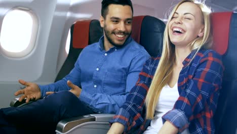 Auf-einem-kommerziellen-Flug-erzählt-Hispanic-Mann-seine-schöne-Blonde-Freundin-lustige-Geschichte.-Beide-lachen.-Sie-Reisen-im-neuen-Flugzeug,-mit-der-Sonne-durch-das-Fenster.