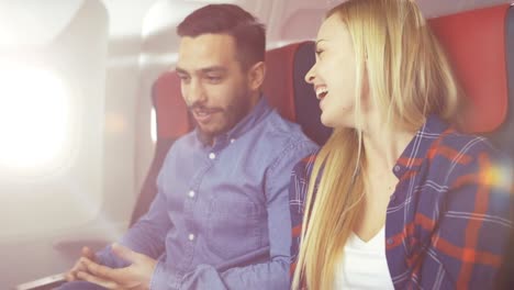 On-a-Commercial-Plane-Flight-Handsome-Hispanic-Man-Tells-Funny-Story-to-His-Beautiful-Blonde-Girlfriend.-Both-Laugh.-They-Travel-in-New-Airplane,-with-Sun-Shining-Through-the-Window.