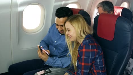 En-un-tablero-de-avión-comercial-hermosa-rubia-joven-con-macho-hispano-Ver-redes-sociales-Smartphone-y-risa.-Pasajero-Senior-lee-el-libro.