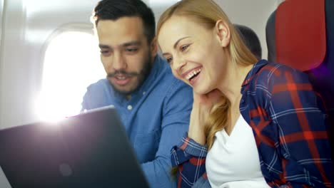 On-a-Board-of-Commercial-Airplane-Beautiful-Young-Blonde-with-Handsome-Hispanic-Male-Watch-Movies-on-a-Laptop-and-Laugh.-Sun-Shines-Through-Aeroplane-Window.