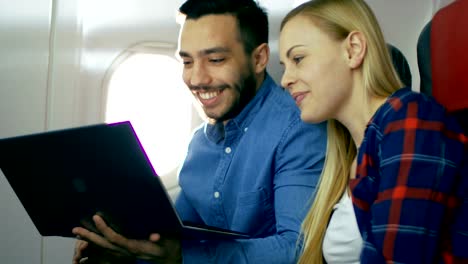 On-a-Board-of-Commercial-Airplane-Beautiful-Young-Blonde-with-Handsome-Hispanic-Male-Watch-Movies-on-a-Laptop-and-Laugh.-Sun-Shines-Through-Aeroplane-Window.