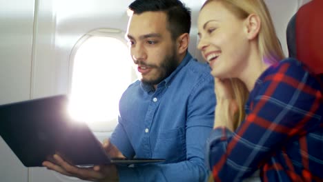 On-a-Board-of-Commercial-Airplane-Beautiful-Young-Blonde-with-Handsome-Hispanic-Male-Watch-Movies-on-a-Laptop-and-Laugh.-Sun-Shines-Through-Aeroplane-Window.