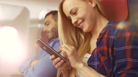 On-Board-of-Commercial-Airplane-Beautiful-Young-Blonde-Uses-Smartphone-while-Her-Hispanic-Male-Neighbor-Sleeps.-Sun-Shines-Through-Plane-Window.