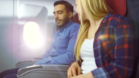 On-a-Plane-Beautiful-Blonde-Female-and-Handsome-Hispanic-Male-Fasten-They're-Seat-Belts-and-Ready-to-Take-off/-Landing.-Sun-Shines-Through-Airplane's-Window.