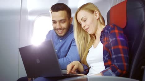 Bei-einem-Board-von-kommerziellen-Flugzeug-schöne-junge-Blondine-mit-schönen-hispanische-männlich-Filme-auf-einem-Laptop-und-lachen.-Durch-Flugzeug-Fenster-scheint-die-Sonne.