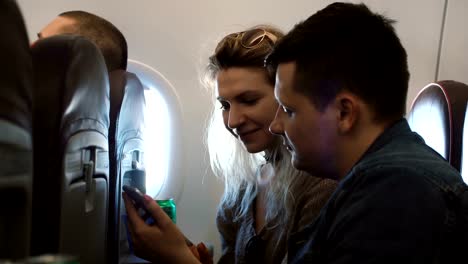 Young-happy-couple-sitting-on-the-plane-near-the-window-and-using-smartphone.-Man-and-woman-traveling-by-airplane