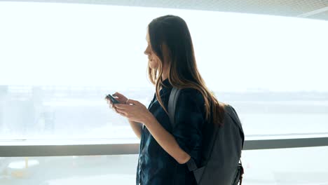 Junges-Mädchen-mit-Smartphone-in-der-Nähe-von-Flughafen-Fenster.-Glücklich-Europäerin-mit-Rucksack-nutzt-mobile-app-im-Terminal.-4K