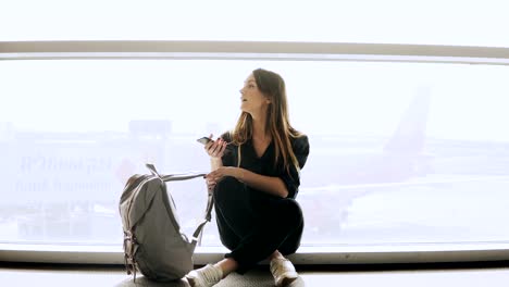 Happy-woman-sits-with-smartphone-by-airport-window.-Caucasian-girl-with-backpack-using-messenger-app-in-terminal.-4K