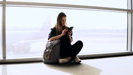 Linda-chica-se-encuentra-usando-el-teléfono-por-la-ventana-de-terminal.-Europeo-femenino-pasajero-con-mochila-escribiendo-y-sonriendo-feliz.-4K