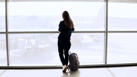 Young-girl-takes-selfie-photo-near-airport-window.-Happy-European-tourist-with-backpack-uses-smartphone-in-terminal.-4K