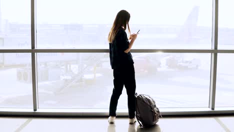 Muchacha-lleva-selfie-junto-a-la-ventana-de-aeropuerto,-aleja.-Caucásica-mujer-turista-feliz-con-smartphone-en-pasillo-de-la-terminal.-4K