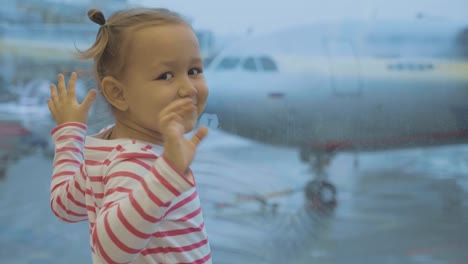 Kleines-Mädchen-steht-neben-Fenster-am-Flughafen-und-Wellen-in-Kamera