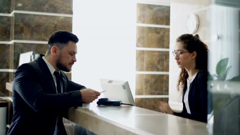 Businessman-guest-talking-with-receptionist-girl-standing-at-desk-in-hotel-reception-and-paying-bill-with-credit-card-via-contactless-payment-technology-.-Business,-travel-and-people-concept