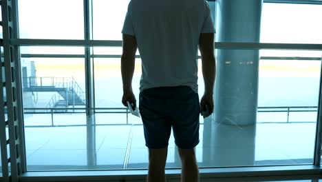 Young-man-with-passport-and-ticket-standing-in-terminal-of-airport-and-looking-at-window.-Handsome-guy-waiting-for-his-airplane.-Rear-back-view-Close-up