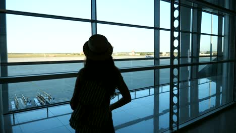 Young-woman-in-hat-with-backpack-coming-to-window-in-terminal-of-airport-and-looking-at-runway.-Girl-waiting-for-her-airplane.-Tourism-and-travel-concept.-Rear-back-view-Close-up