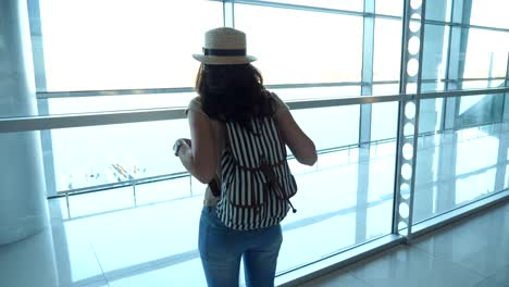 Young-woman-in-hat-with-backpack-standing-in-terminal-of-airport-and-looking-at-window.-Girl-waiting-for-her-airplane-and-then-leaves.-Tourism-and-travel-concept.-Rear-back-view-Close-up