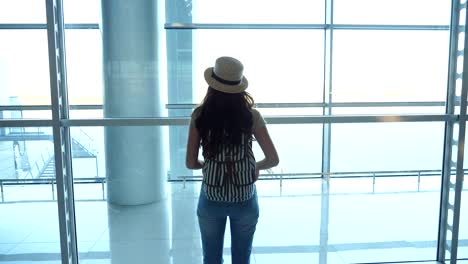 Young-woman-in-hat-with-backpack-coming-to-window-in-terminal-of-airport-and-looking-at-runway.-Girl-waiting-for-her-airplane.-Tourism-and-travel-concept.-Rear-back-view-Close-up