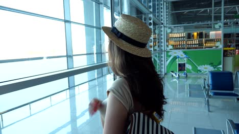 Young-woman-in-hat-with-backpack-standing-in-terminal-of-airport-and-looking-at-window.-Brown-haired-girl-waiting-for-her-airplane.-Tourism-and-travel-concept.-Close-up-Side-view