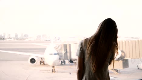 Young-European-girl-talks-on-smartphone-and-smiles,-ends-call-at-airport-terminal-window,-airplane-in-the-background