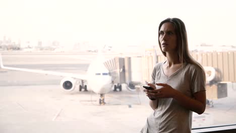 Young-European-woman-standing-near-airport-terminal-window-gets-upset-and-frustrated-looking-at-phone-and-smart-watch