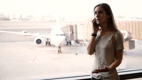 Young-European-woman-talking-on-the-phone-near-airport-terminal-window-upset-and-frustrated-after-missing-flight