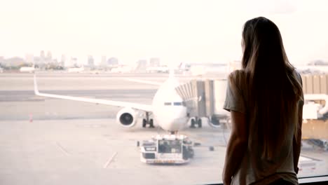 Beautiful-happy-Caucasian-girl-with-long-hair-walks-up-to-airport-terminal-window-to-enjoy-the-view-of-airplanes