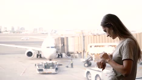 Joven-empresaria-caucásicas-de-pie-en-la-ventana-de-salón-terminal-Aeropuerto-usando-reloj-elegante,-mirando-aviones