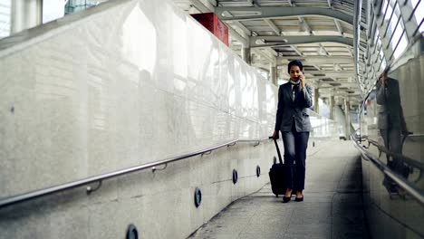 Asian-business-woman-talking-the-mobile-phone-while-dragging-a-wheeled-luggage-at-the-airport-ramp-for-business-trip