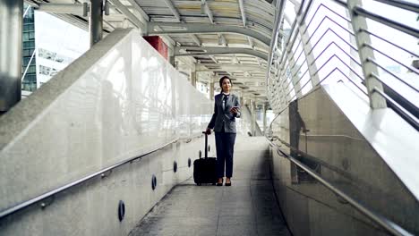 Asian-business-woman-talking-the-mobile-phone-while-dragging-a-wheeled-luggage-at-the-airport-ramp-for-business-trip