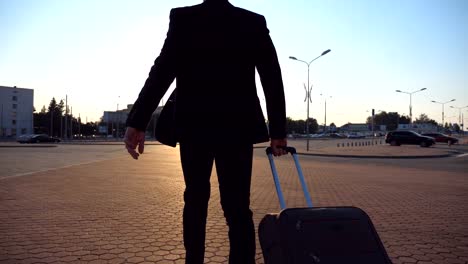 Unrecognizable-businessman-in-black-suit-is-late-for-the-flight-and-quickly-going-with-his-suitcase-to-airport.-Young-man-looking-at-his-watch-and-in-hurry-walking-with-luggage-to-terminal.-Rear-view