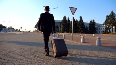 Dolly-la-foto-de-la-joven-confía-en-un-formal-traje-negro-caminando-con-su-equipaje-en-calle-urbana.-Próspero-hombre-de-negocios-va-a-la-terminal-del-aeropuerto-y-tirando-de-maleta-sobre-ruedas-al-atardecer.-Cierre-para-arriba