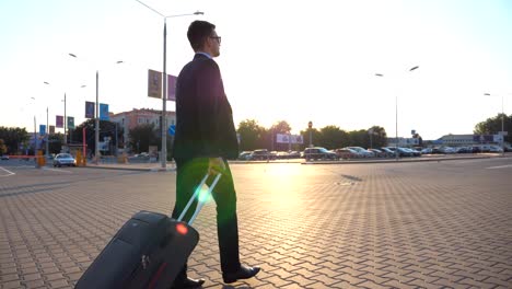 Dolly-shot-of-confident-young-man-in-a-formal-black-suit-walking-with-his-luggage-on-urban-street.-Successful-businessman-going-to-airport-terminal-and-pulling-suitcase-on-wheels-at-sunset.-Close-up