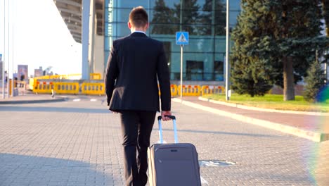 Rear-view-of-unrecognizable-young-businessman-walking-to-airport-terminal-and-pulling-suitcase-on-wheels-at-sunset.-Successful-male-business-person-going-with-his-luggage-on-city-street.-Close-up