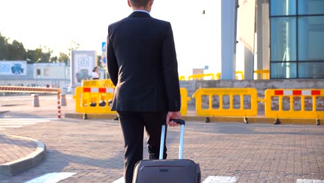 Dolly-shot-of-confident-young-man-in-a-formal-black-suit-walking-with-his-luggage-on-urban-street.-Successful-businessman-going-to-airport-terminal-and-pulling-suitcase-on-wheels-at-sunset.-Close-up