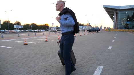 Happy-successful-businessman-walking-on-city-street-and-pulling-suitcase-on-wheels-after-flight-at-sunset.-Young-man-going-with-a-jacket-on-his-shoulder-against-the-background-of-airport.-Close-up