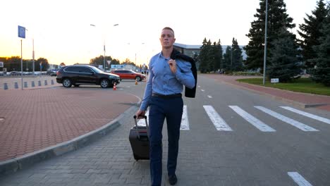 Young-man-walking-on-urban-environment-with-a-jacket-on-his-shoulder-and-pulling-suitcase-on-wheels.-Successful-businessman-going-with-luggage-from-the-airport-on-city-street.-Close-up-Dolly-shot