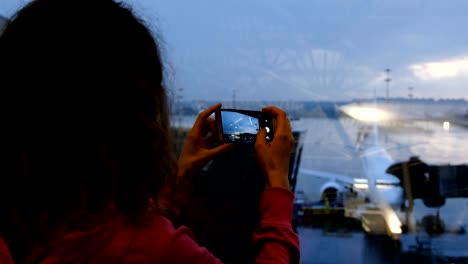 Frau-in-einem-Flughafen-Wartezimmer-nimmt-ein-Bild-von-einem-Flugzeug-durch-einen-Spiegel,-eine-Smartphone-Nahaufnahme