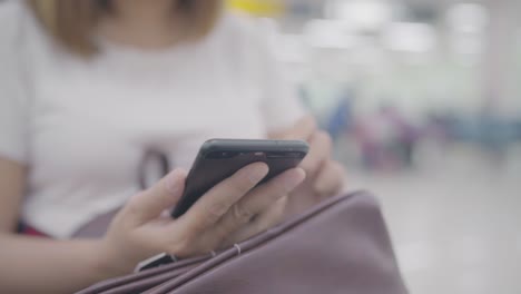 Gerne-Asiatin-mit-und-überprüfen-ihr-Smartphone-sitzend-auf-Stuhl-im-terminal-Halle-während-des-Wartens-ihr-Fluges-am-Abfluggate-im-internationalen-Flughafen.-Frauen-glücklich-im-Flughafenkonzept.