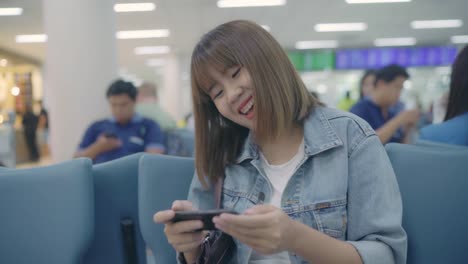 Feliz-mujer-asiática-utilizando-y-controlando-su-smartphone-sentado-en-silla-en-la-sala-terminal-mientras-espera-su-vuelo-en-la-puerta-de-salida-en-el-aeropuerto-internacional.-Mujer-feliz-en-concepto-de-aeropuerto.