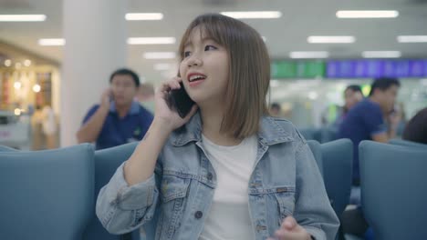 Happy-Asian-woman-using-and-checking-her-smartphone-while-sitting-on-chair-in-terminal-hall-while-waiting-her-flight-at-the-departure-gate-in-international-airport.-Women-happy-in-airport-concept.